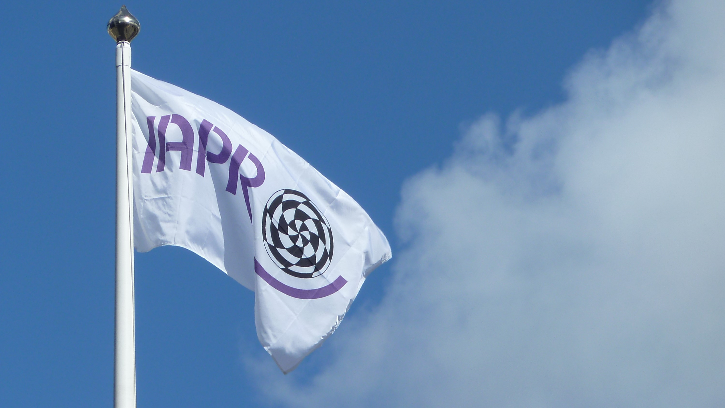 A white flag with the IAPR logo flying in front of a blue sky and white clouds.